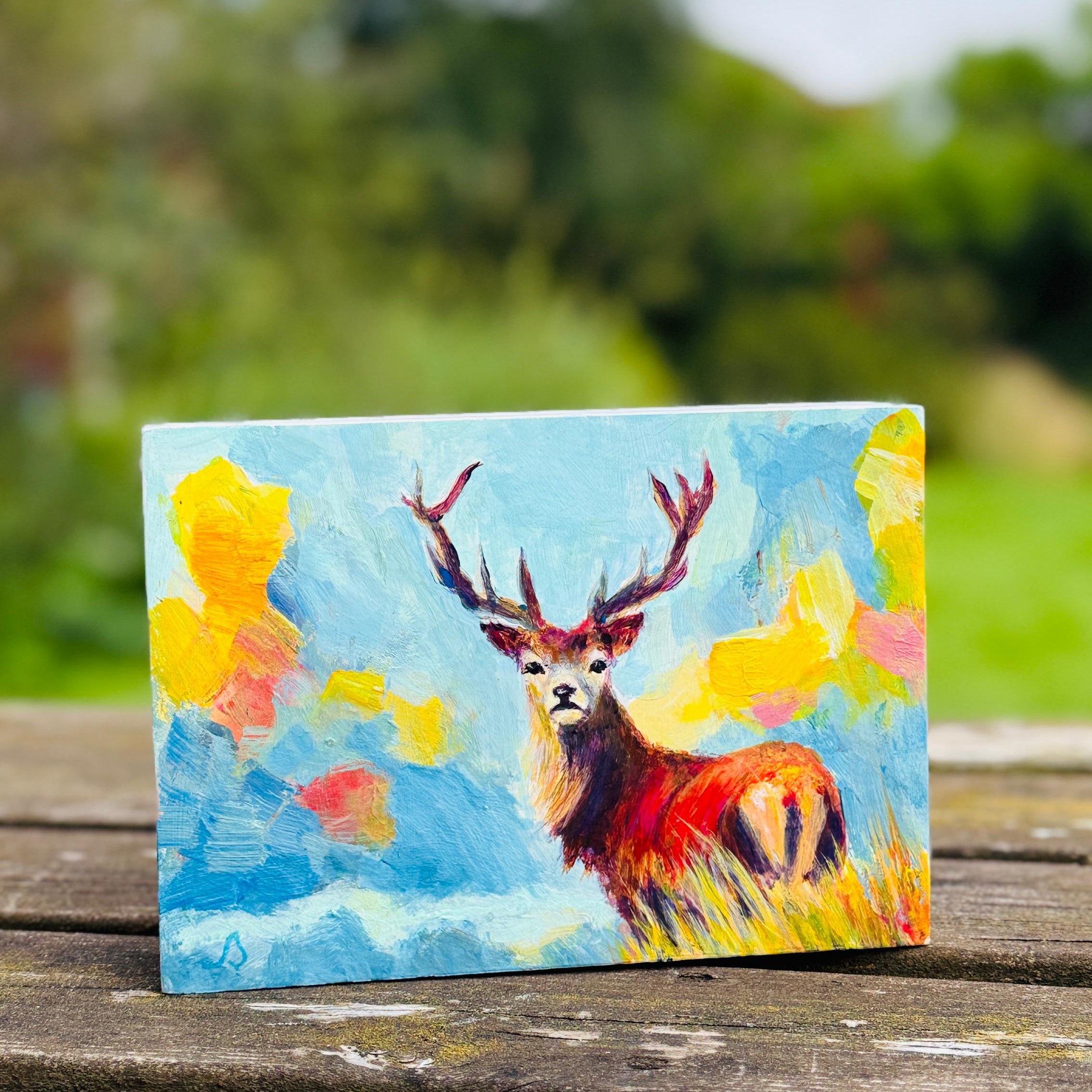 Hand holding a small painting of a stag deer with a colorful expressive landscape background
