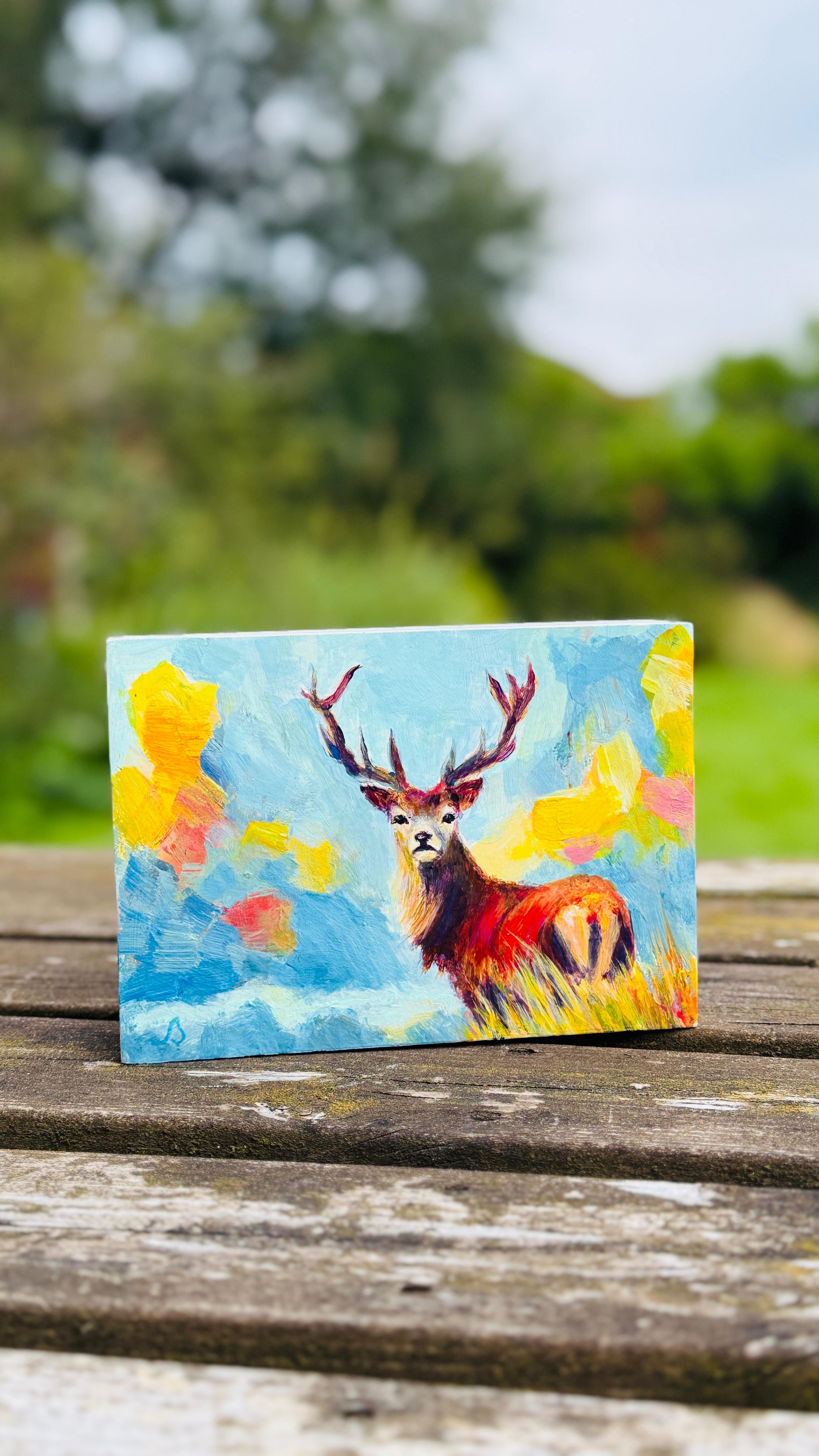 Hand holding a small painting of a stag deer with a colorful expressive landscape background