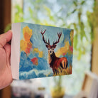 Hand holding a small painting of a stag deer with a colorful expressive landscape background