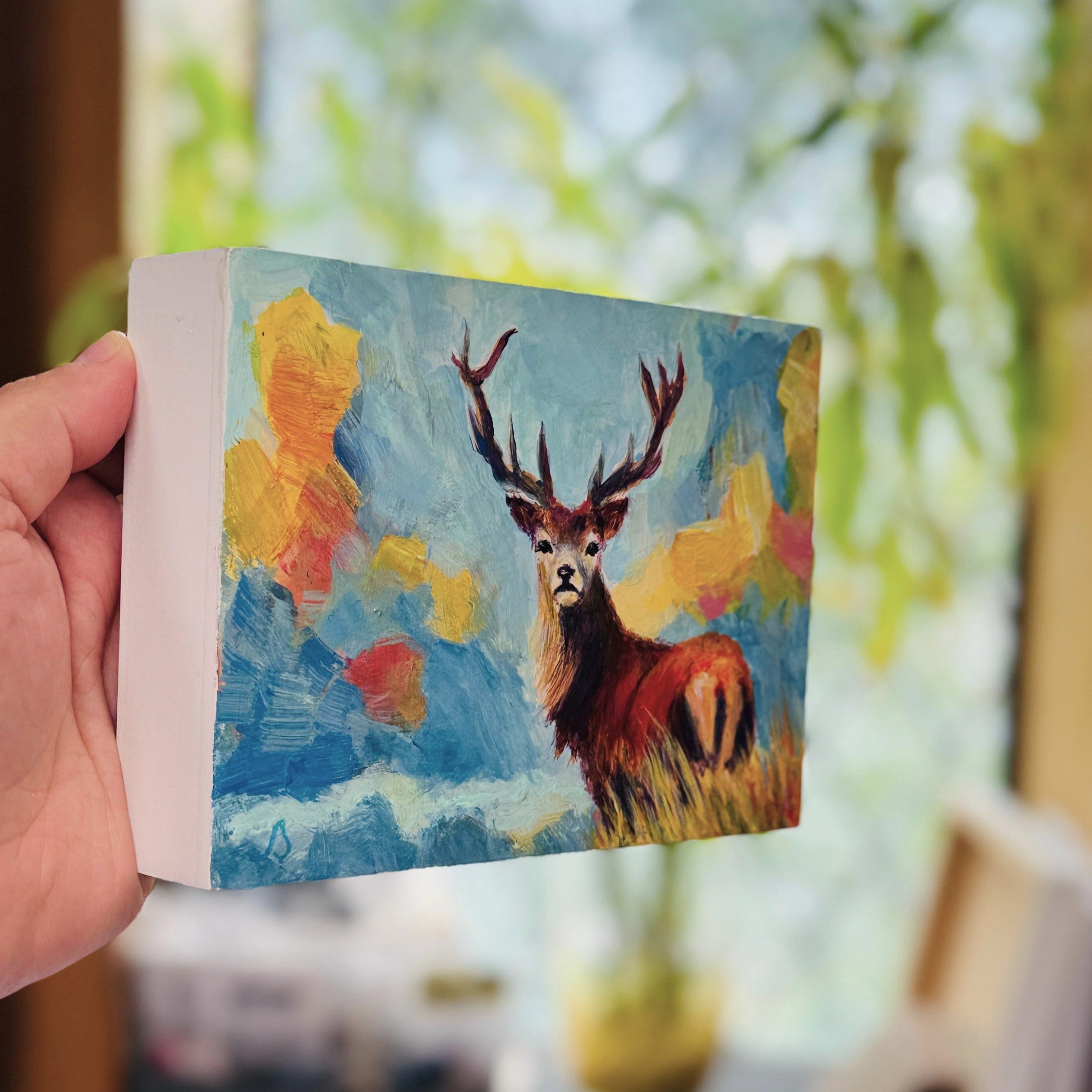 Hand holding a small painting of a stag deer with a colorful expressive landscape background